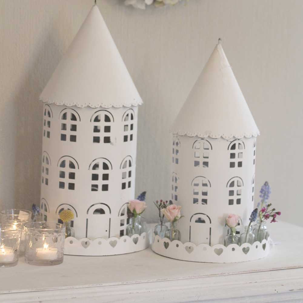 Zwei weiße, dekorative VL Home - Vogelhäuser rund mit Glasflaschen in Turmform mit Herzausschnitt stehen auf einem Tisch. Daneben stehen drei kleine brennende Kerzen.
