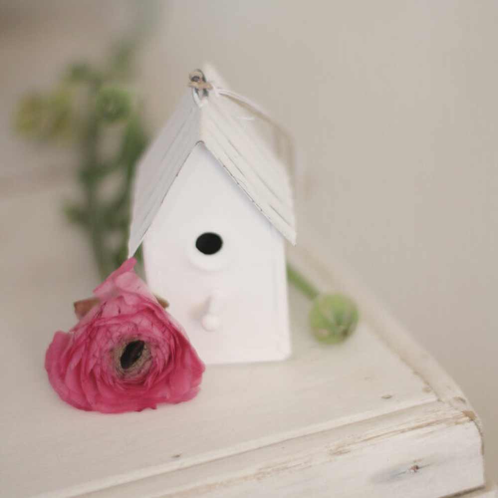 Das VL Home – Vogelhaus schmal weiß aus Metall steht auf einer hellen Fläche neben einer rosa Blüte und grünen Knospen.