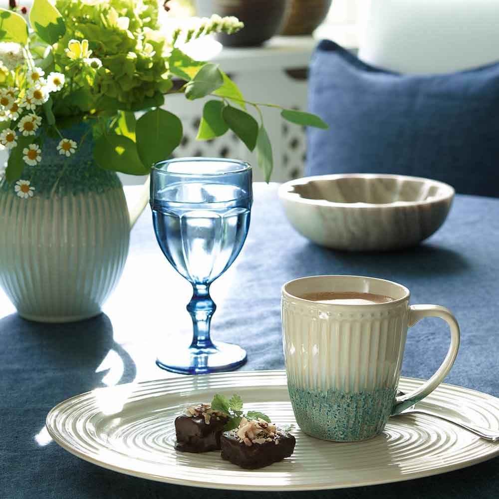 Eine gemütliche Wohnatmosphäre mit einem Tablett mit einer GreenGate - Alice Latte Tasse, einem Glas Wasser und Schokoladengebäck neben einer Vase mit frischen Blumen.