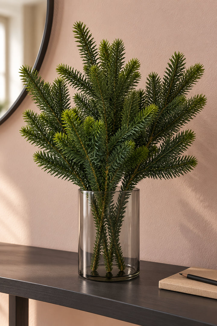 Drei künstliche Kiefernzweige ohne Zapfen in einer rauchgrauen Glasvase vor einer rosafarbenen Wand.