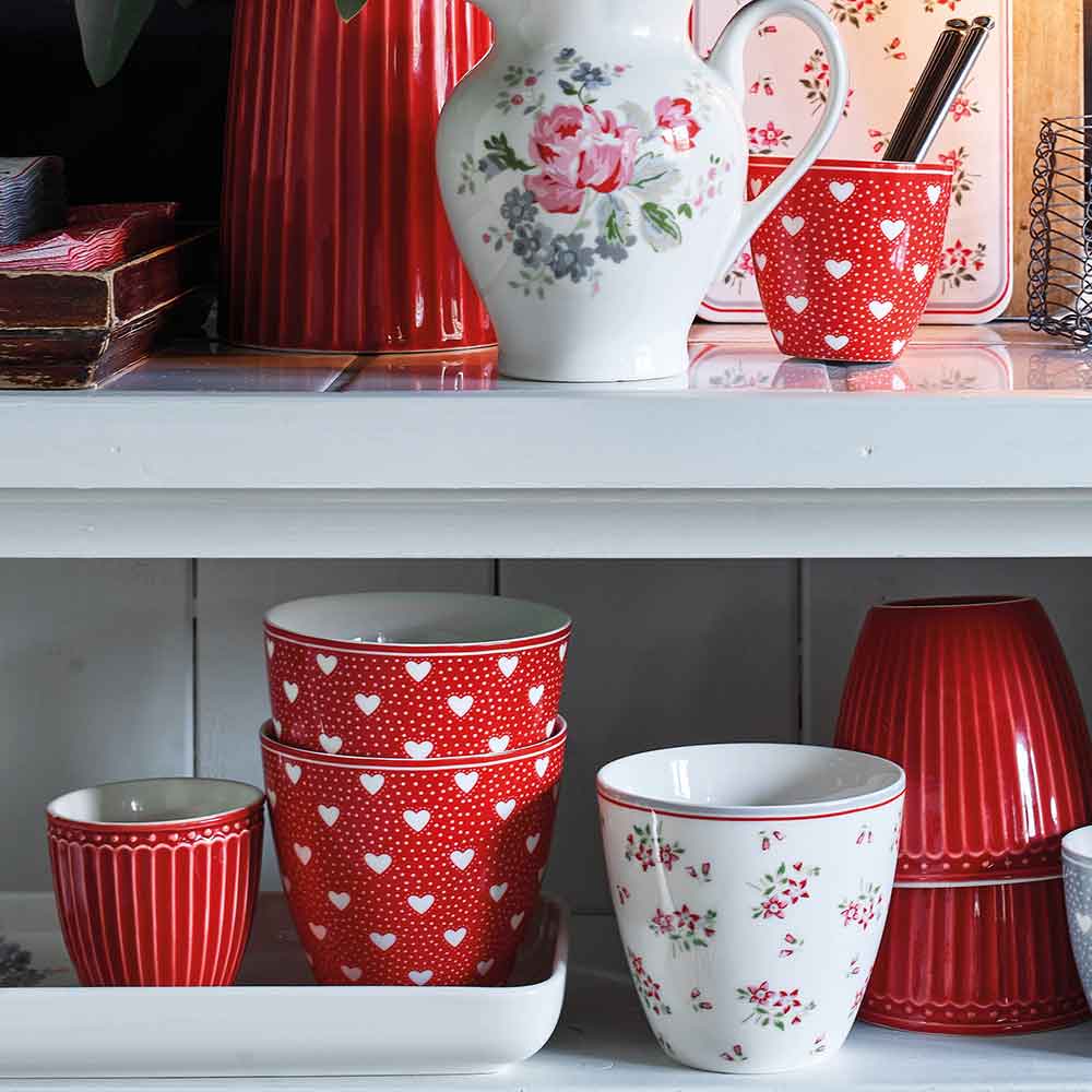 Eine Kollektion von GreenGate - Alice Mini Latte Cups in Rot und Weiß aus Keramikküchengeschirr auf Regalen, darunter gemusterte Becher, Krüge und Tassen mit Herz- und Blumendesigns.