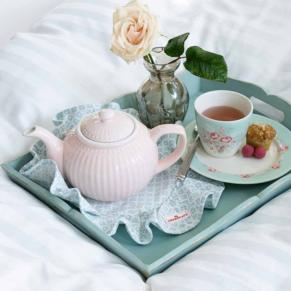 Ein gemütliches Tee-Set auf einem Bett mit einer GreenGate – Alice Teekanne, einer Tasse Tee, Süßigkeiten und einer Rose in einer Vase auf einem blauen Tablett.