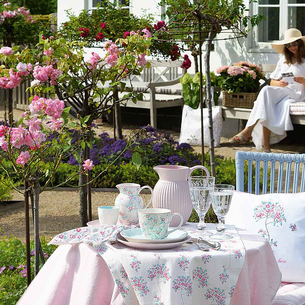 Eine gemütliche Tee-Session im Freien mit dem GreenGate - Allyna Geschirrtuch White als Tischdecke, geschmückt mit Teetassen und einem rosa Krug. Blühende Bäume sorgen für eine ruhige Kulisse, während eine Person mit Sonnenhut auf einer Bank in der Nähe entspannt.