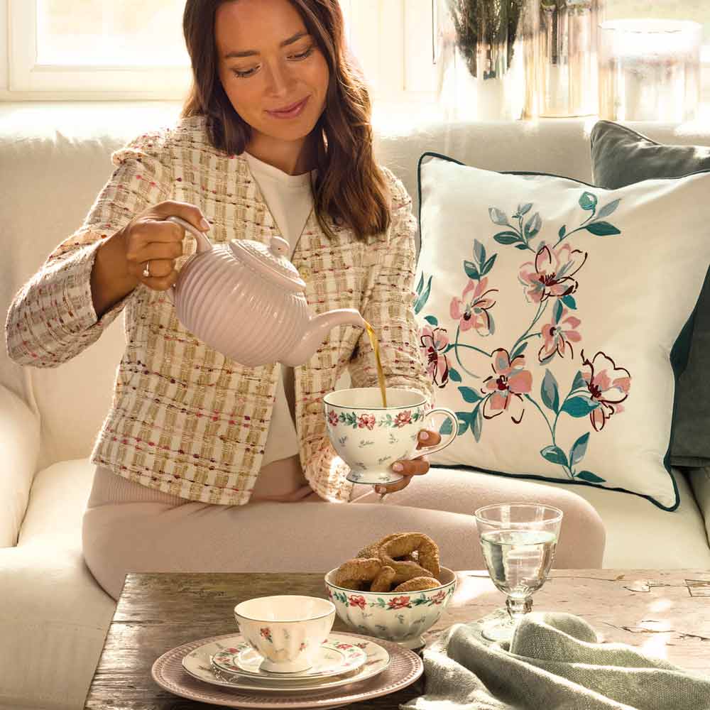 Eine Frau gießt Tee aus einer Teekanne in einen weißen GreenGate - Jalia Eierbecher und sitzt auf einem Sofa mit dekorativen Kissen mit Blumenmuster. Eine Tasse, eine Schüssel und ein Glas Wasser stehen auf dem Holztisch vor ihr.
