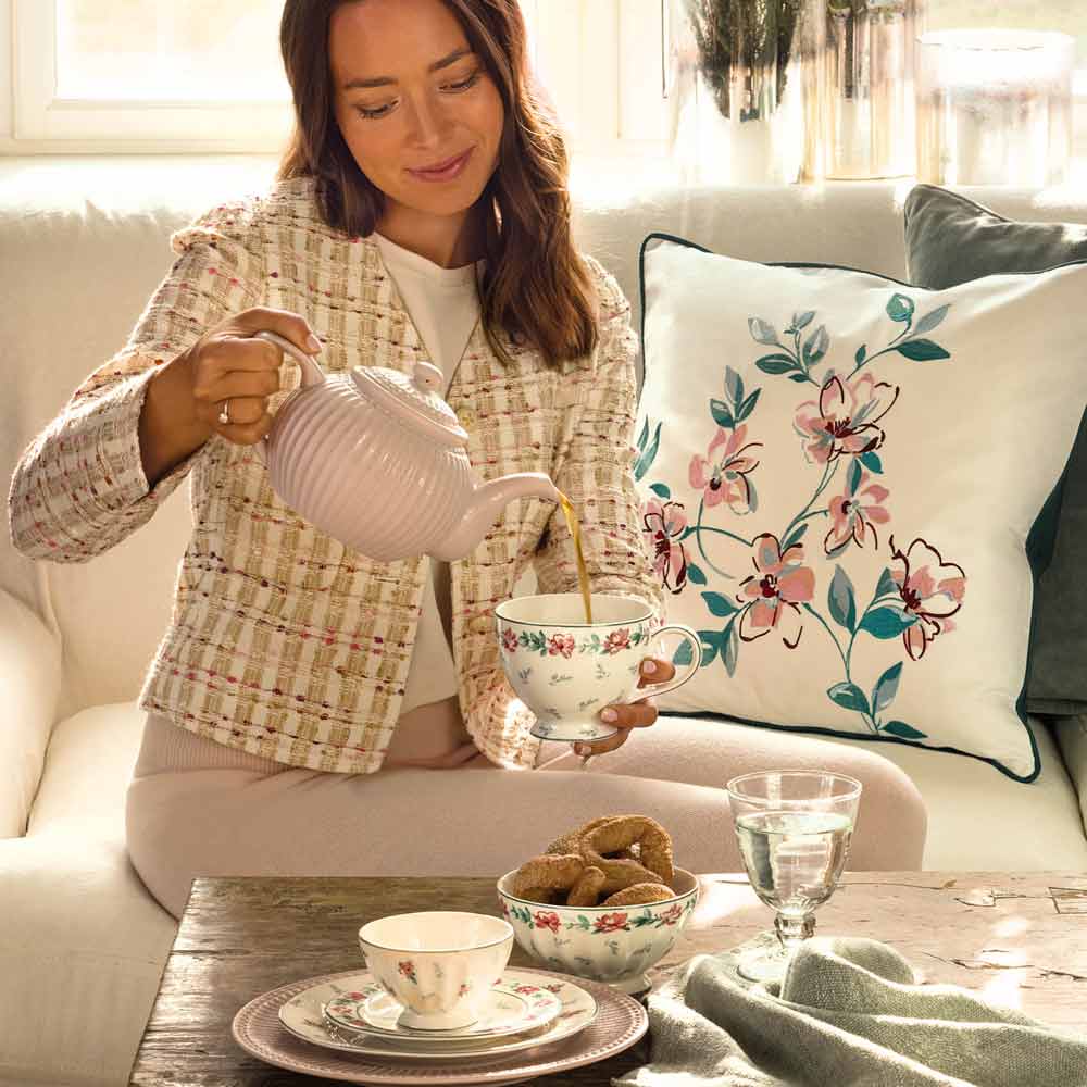 Eine Frau gießt Tee aus einer Teekanne in einen weißen GreenGate - Jalia Eierbecher und sitzt auf einem Sofa mit dekorativen Kissen mit Blumenmuster. Eine Tasse, eine Schüssel und ein Glas Wasser stehen auf dem Holztisch vor ihr.