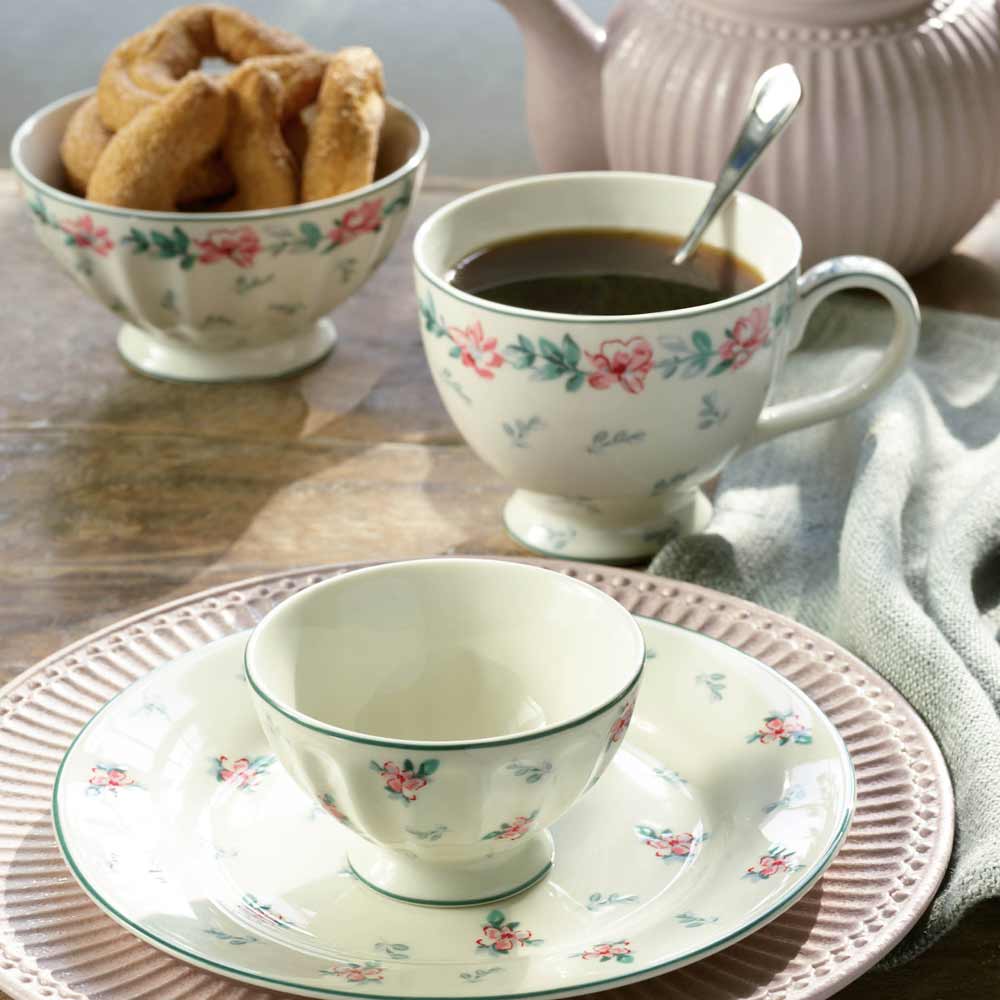 Ein weißer GreenGate - Jalia Eierbecher, eine kleine Schüssel und ein Teller mit Blumenmuster sind auf einem Tisch neben einer Tasse schwarzen Kaffee, einer Schüssel Donuts und einer Teekanne arrangiert.