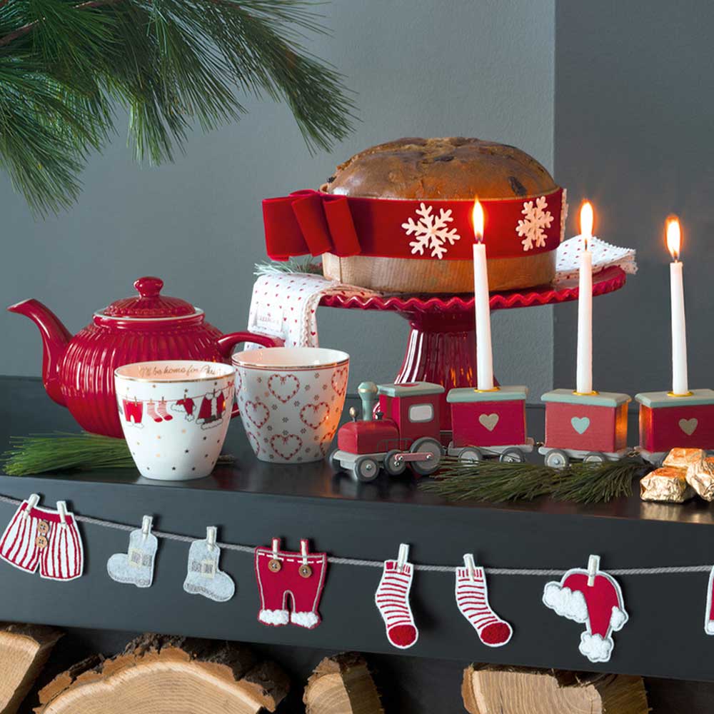 Eine festliche Weihnachtsdekoration mit einer GreenGate - Alice Teekanne, einem Becher, einem Panettone auf einem Ständer und einer Spielzeugeisenbahn, geschmückt mit brennenden Kerzen auf einem Regal über Brennholz.