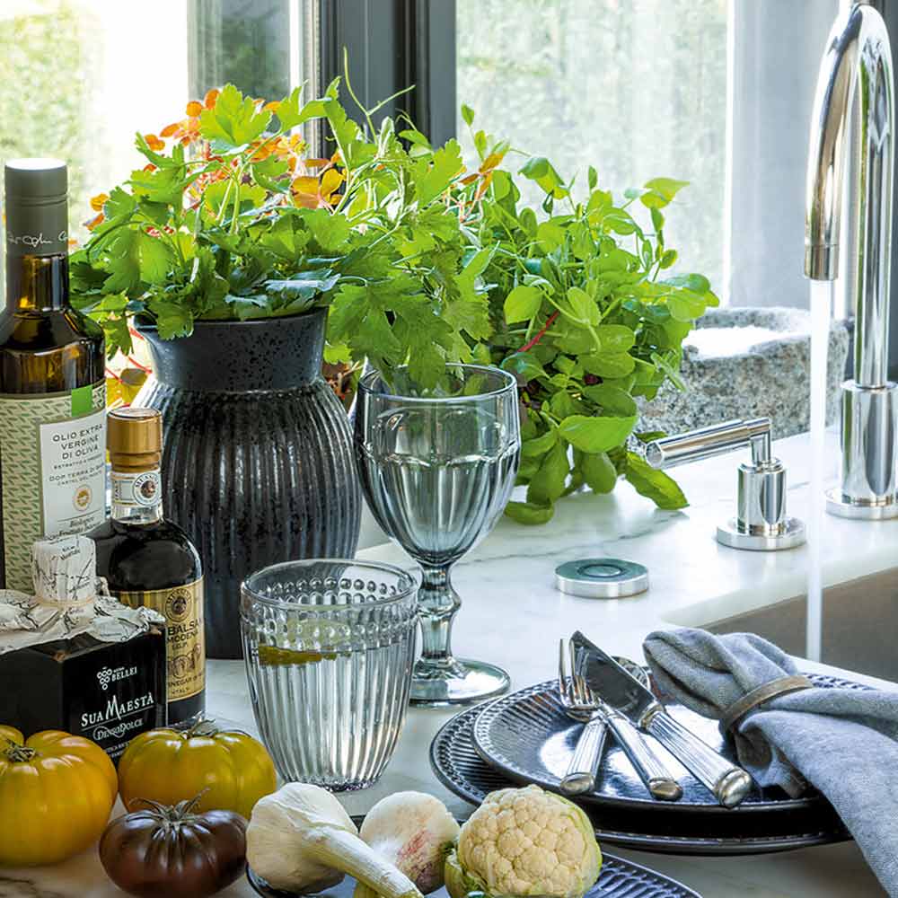 Eine Küchentheke, dekoriert mit frischen Kräutern, Gemüse, Olivenöl und elegantem Glasgeschirr neben einer Spüle mit Blick aus dem Fenster. GreenGate - Alice Latte Cup.