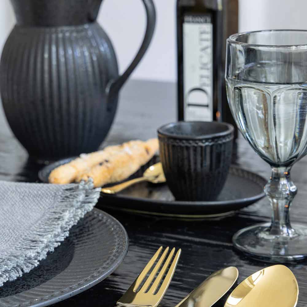 Elegantes Esszimmer-Setup mit schwarzem Keramikgeschirr, goldenem Besteck und einer durchsichtigen GreenGate - Alice Latte Cup, mit einem dezenten Hintergrund aus einer dunklen Flasche und einer strukturierten grauen Serviette.