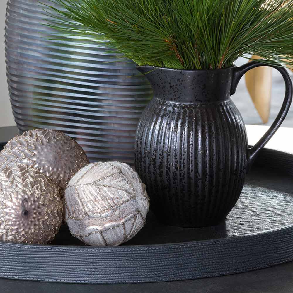 Eine schwarze Keramikvase mit strukturierter Oberfläche hält Tannenzweige, neben zwei dekorativen glitzernden Kugeln auf einem grauen Tablett.