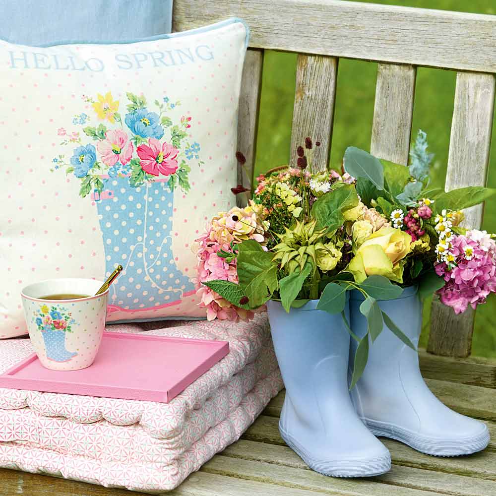 Frühlingsthema im Freien mit einem dekorierten Kissen, einem GreenGate - Josefina Latte Cup Summer White, einem Buch und mit Blumen gefüllten Gummistiefeln auf einer Holzbank.