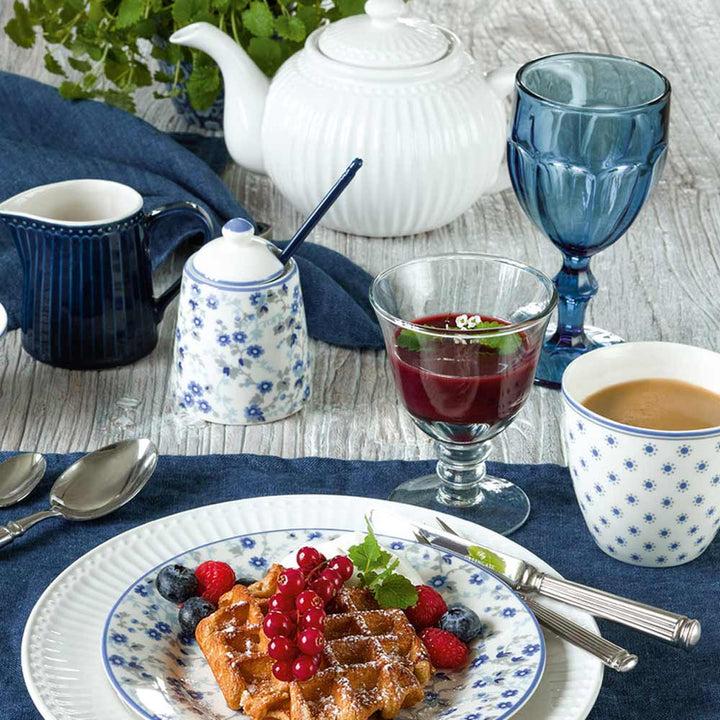 Ein Frühstücksbuffet mit Waffeln mit Beeren, einer Tasse Kaffee und einer GreenGate – Alice Teekanne, serviert auf einem Holztisch mit elegantem blau-weißem Geschirr.