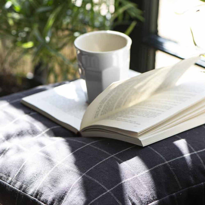 Ein offenes Buch und ein Ib Laursen - Cafe Latte Becher Mynte auf einem karierten Tuch neben einem sonnigen Fenster, mit Pflanzen im Hintergrund.