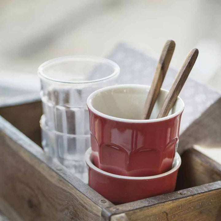Zwei Ib Laursen - Cafe Latte Becher Mynte, einer rot und einer weiß, mit Holzlöffeln darin, auf einem Holztablett mit einem Glas Wasser daneben.