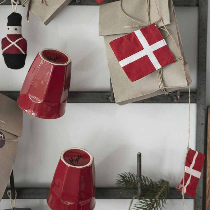Rote Ib Laursen - Cafe Latte Becher Mynte-Becher, die kopfüber an Schnüren hängen, einer mit der Aufschrift „Vega“, neben einer Wand mit einem Dekor mit der dänischen Flagge und einem Stoffkalender.
