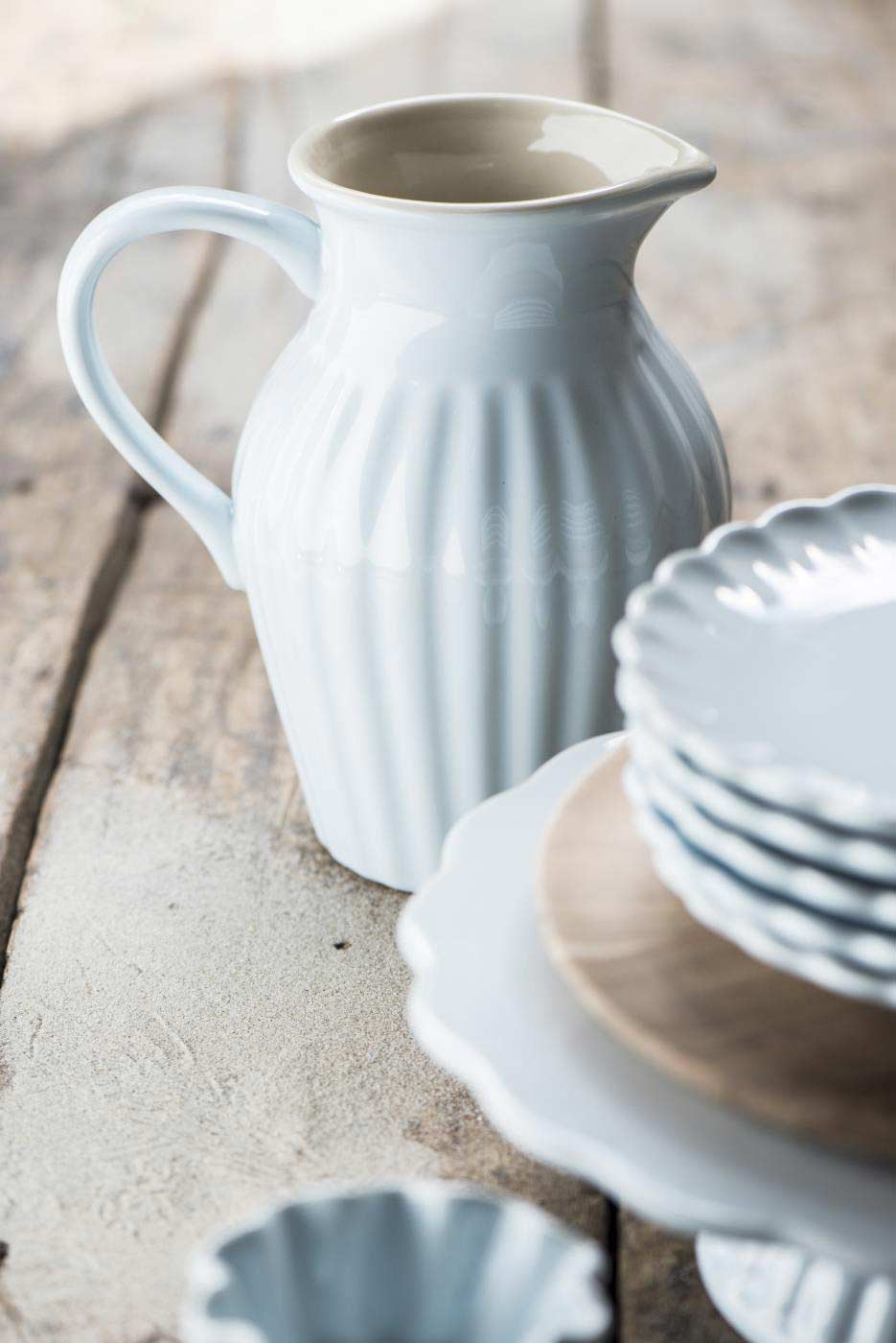 Ein Ib Laursen - Krug Mynte 1,7 Liter mit einem Stapel Teller auf einem rustikalen Holztisch, beleuchtet von sanftem Licht.