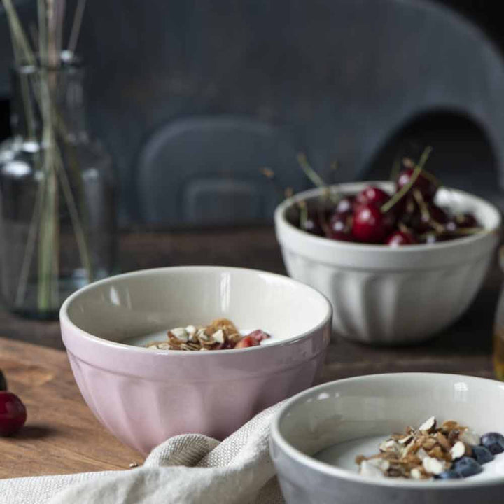 Zwei Schüsseln Joghurt mit Granola und Früchten, eine rosa und eine weiße, in der Ib Laursen - Müslischale Mynte auf einem Holztisch mit einer Schale Kirschen im Hintergrund.