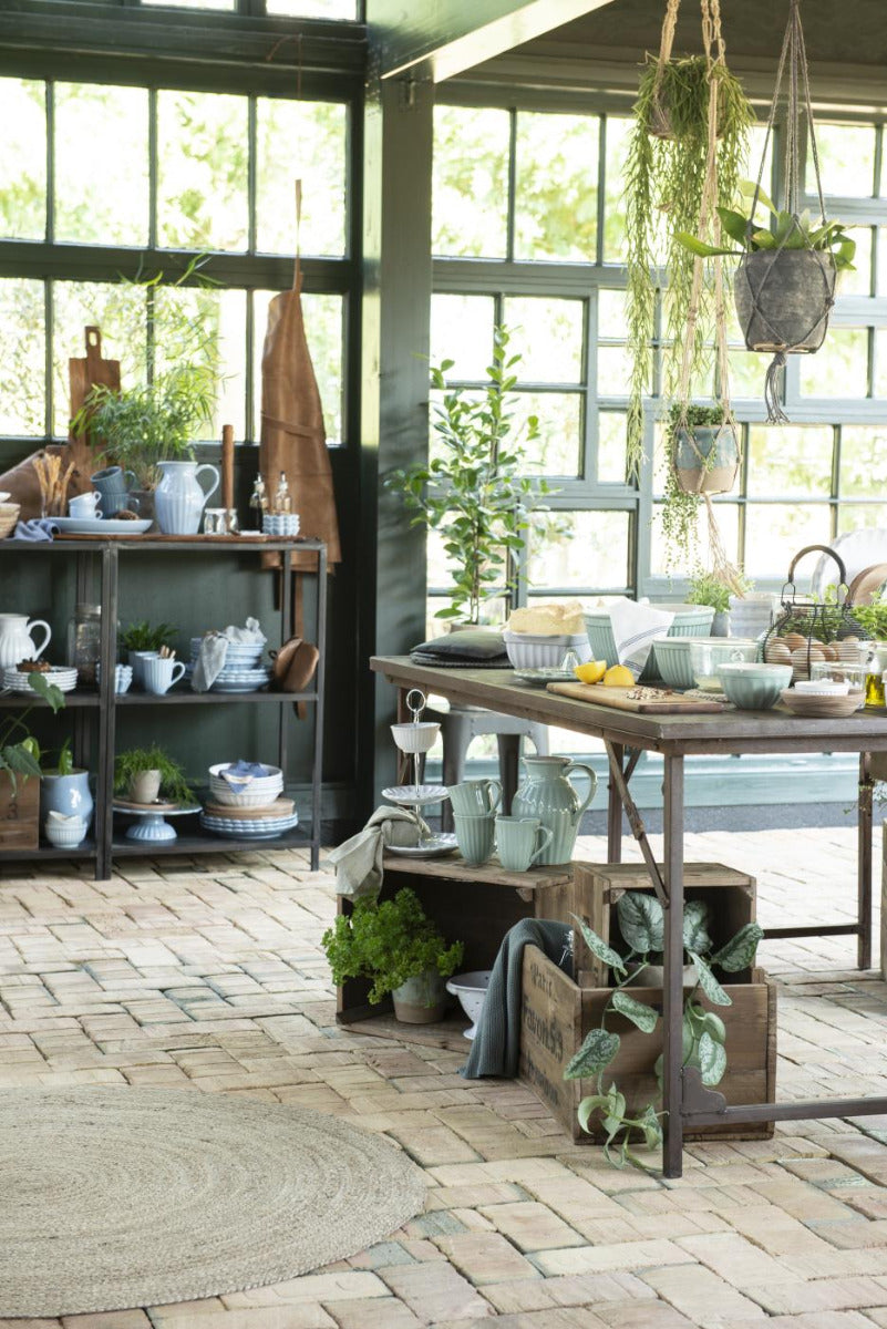 Die Inneneinrichtung eines rustikalen Gartengeschäfts besteht aus einem Holztisch mit Ib Laursen - Krug Mynte 1,7 Liter, Regalen voller Pflanzen und hängenden Töpfen neben großen Fenstern.