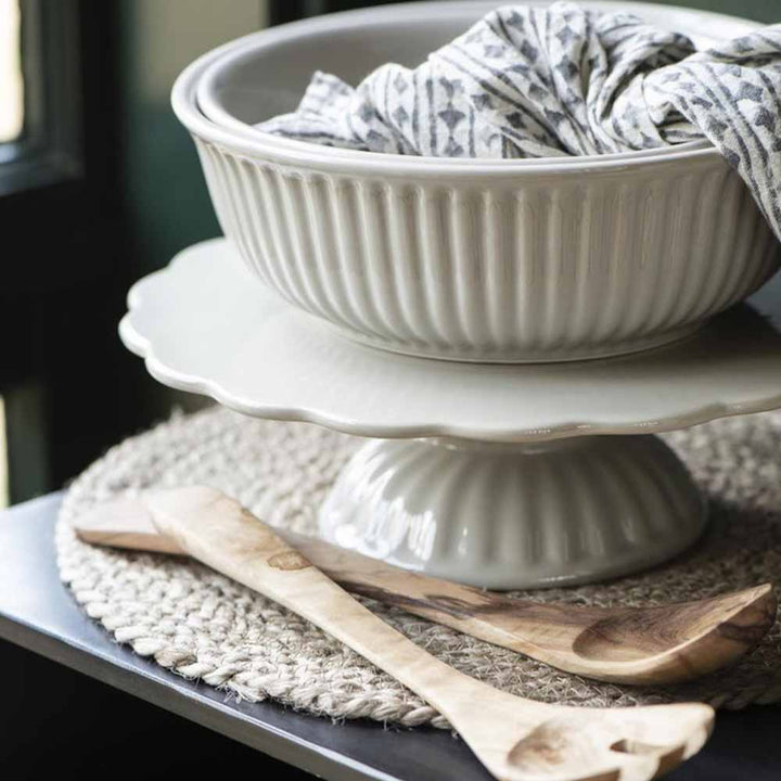 Weiße Keramik Ib Laursen – Tortenplatte Mynte und Teller auf einem Kuchenständer, begleitet von Holzutensilien, auf einer gewebten Matte.