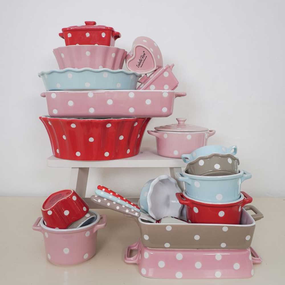 Stapel farbenfroher Isabelle Rose – Tarteform mit Punkten im Tupfenmuster, darunter Töpfe, Pfannen und Utensilien auf weißem Hintergrund.