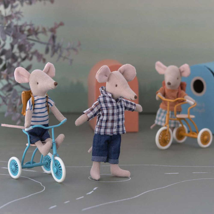 Drei Maileg-Spielzeugmäuse in verspielter Anordnung, eine fährt Dreirad, eine winkend und eine auf einem Roller, alle auf einer gezeichneten Straße mit Hintergrundelementen.