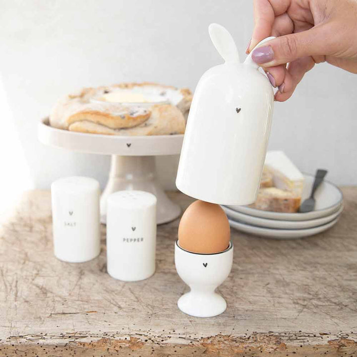 Eine Person hält einen Bastion Collections - Eierwärmer Keramik Hase weiß und einen Hasen auf einem Tisch.