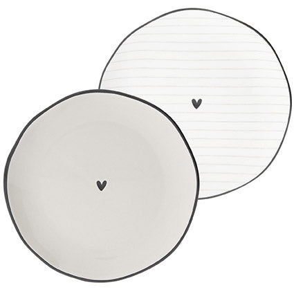 Zwei Bastion-Kollektionen – Keksteller Heart mit schwarzem Rand Beige mit minimalistischem Herzdesign, eine davon mit horizontalen Linien.