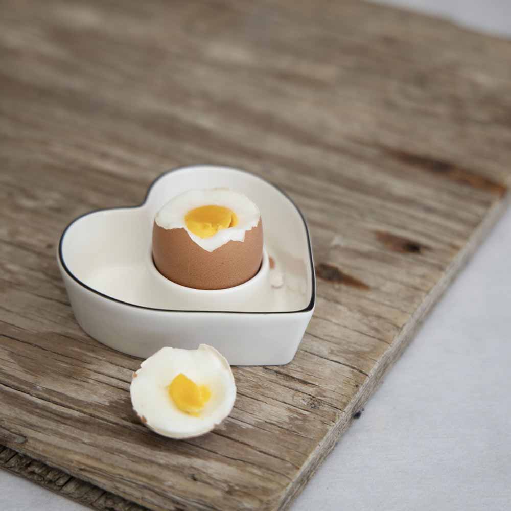 An Bastion Collections – Eierbecherhalter aus Keramik in Herzform in einer herzförmigen Schale auf einem Holztisch.