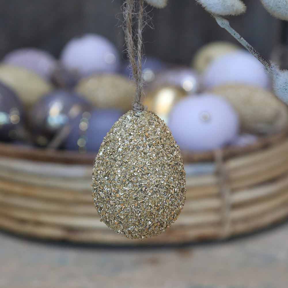 Eine Chic Antique - Osterei mit Goldglitter hängt vor einem Korb gefüllt mit verschiedenen pastellfarbenen Eiern.
