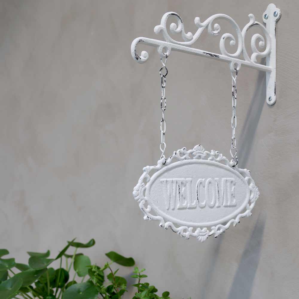 Ein Chic Antique - Schild „Welcome“ hängt an einer dekorativen Metallhalterung an einer hellgrauen Wand, mit grünen Pflanzen in der unteren linken Ecke.