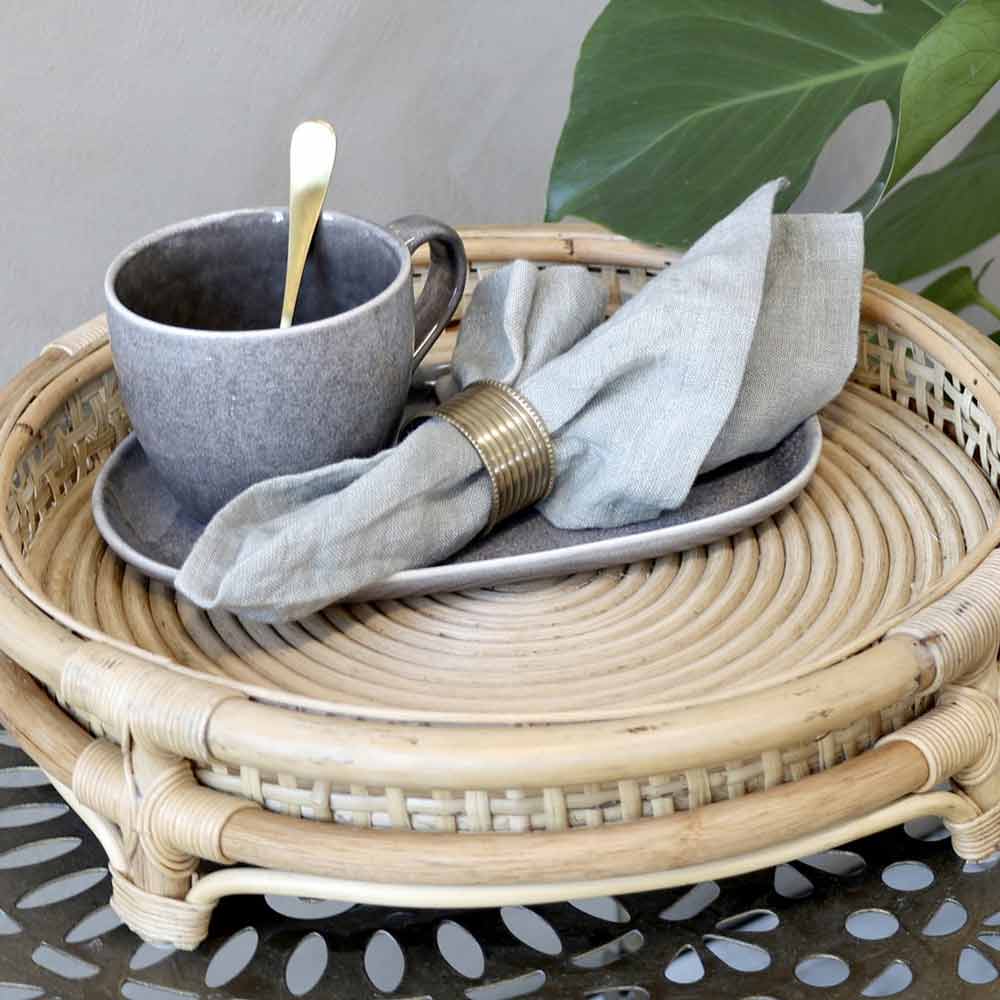 Ein Chic Antique - Tablett, französisch geflochten auf Fuß mit einem Löffel darin, auf einer Untertasse neben einer gefalteten Serviette platziert, alles auf einem Korbtablett auf einem gemusterten Tisch.