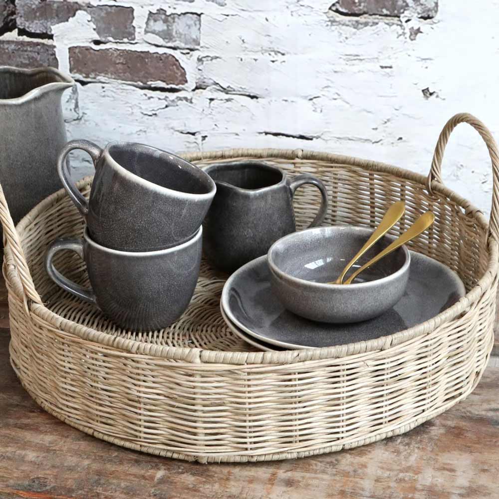 Ein Chic Antique Tablett geflochten mit Griffen natur mit einem Set aus grauem Keramikgeschirr und Bechern sowie Holzbesteck vor einer weißen Ziegelwand.