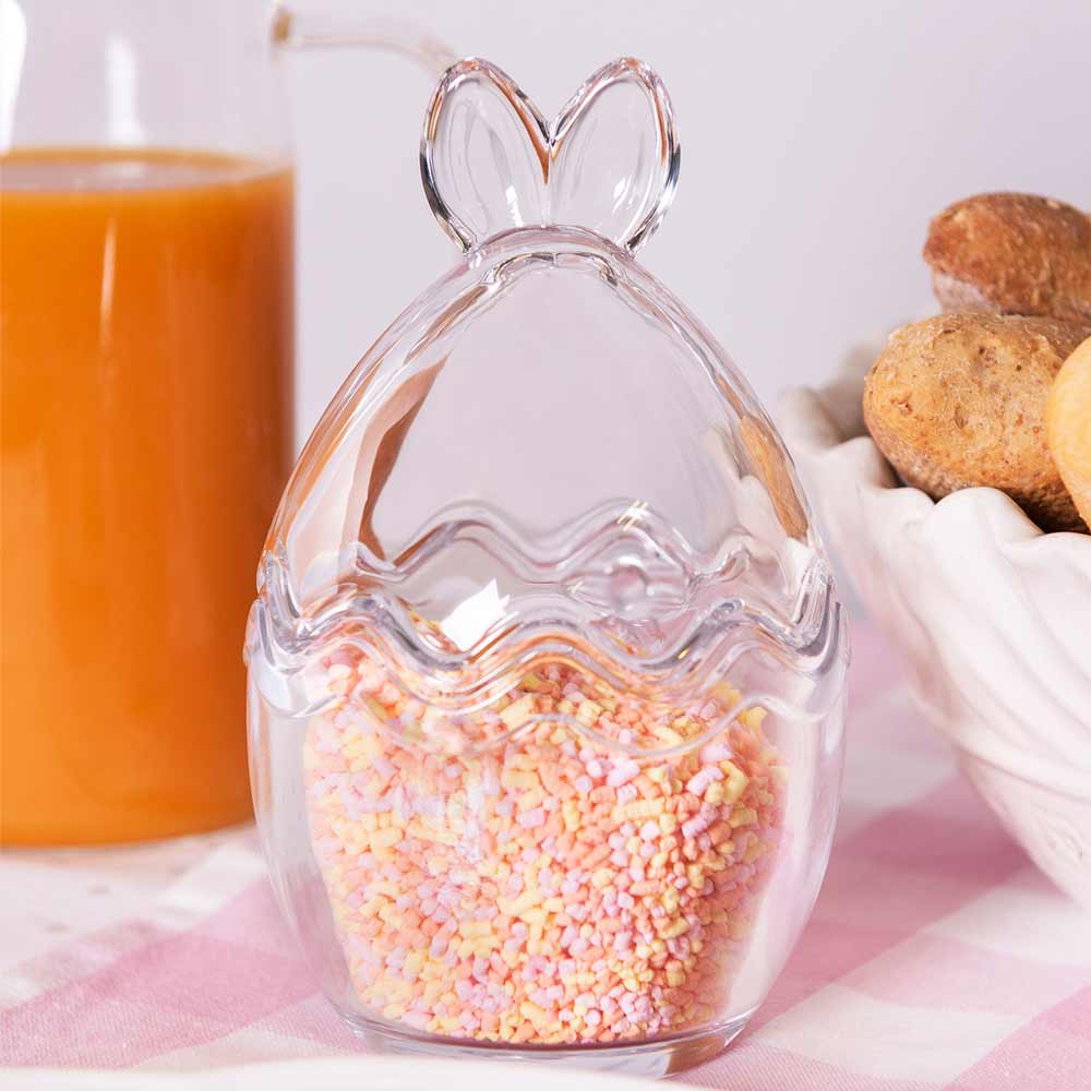 Klarer Glasbehälter in Ostereiform, gefüllt mit bunten Streuseln, mit Keksen und einer Saftflasche im Hintergrund.