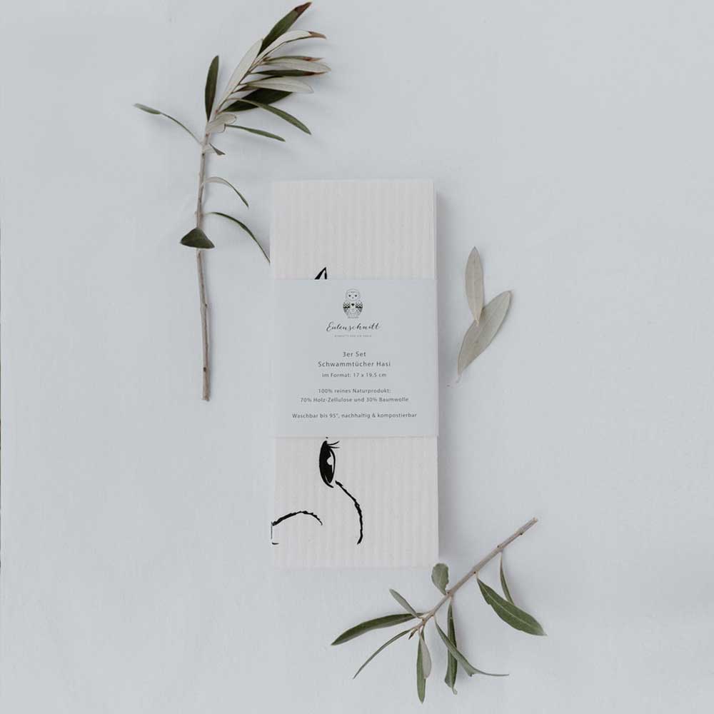 Ein minimalistisches Eulenschnitt - Schwammtücher Hasi 3er Set flankiert von zarten Olivenzweigen auf neutralem Hintergrund.
