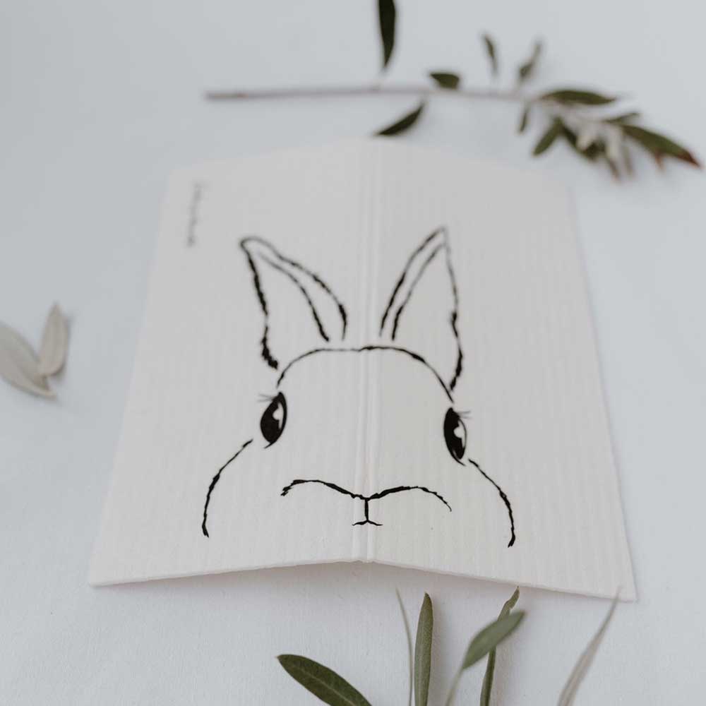 Eine einfache Skizze des Gesichts von Eulenschnitt - Schwammtücher Hasi 3er Set auf einem aufgeschlagenen Notizbuch mit einem Zweig Blätter im Hintergrund.