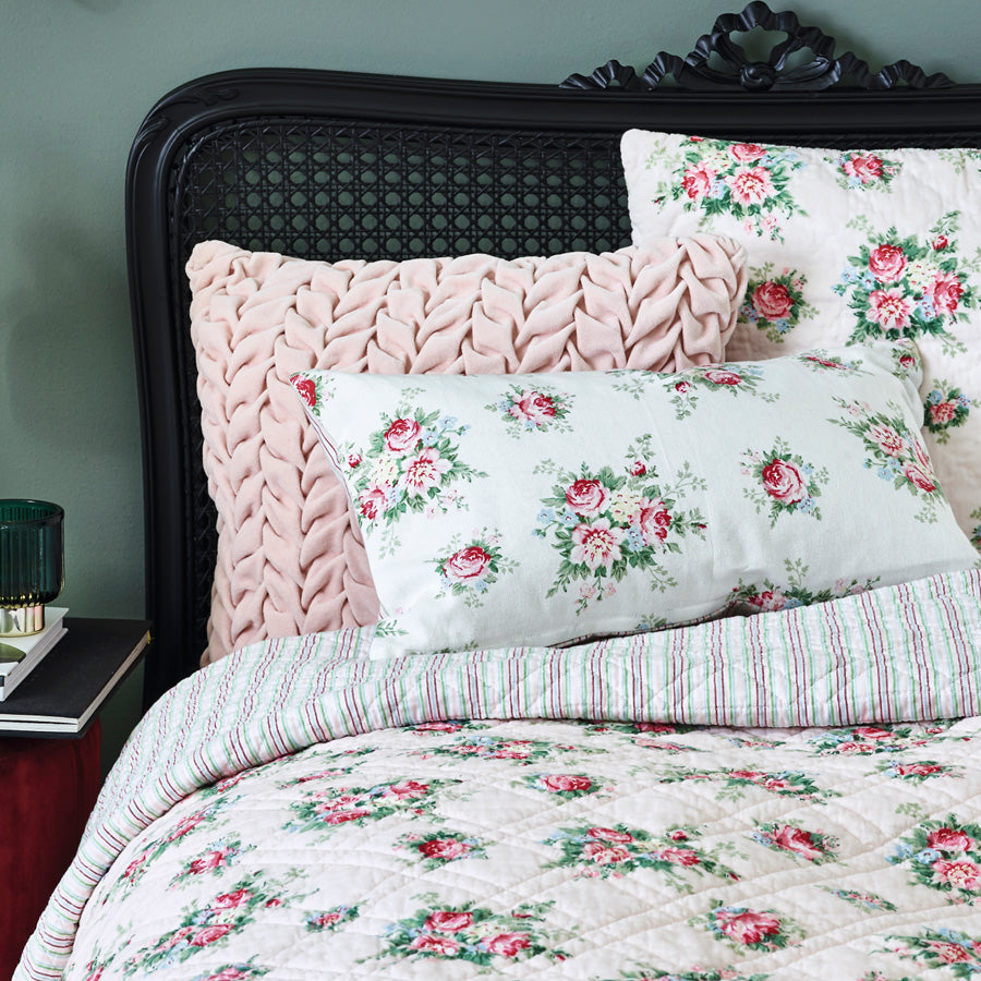 Bett mit dunklem Geflechtkopfteil, einfarbigem rosa Kissen, Rosenmustern und einer umgeschlagenen gestreiften Decke