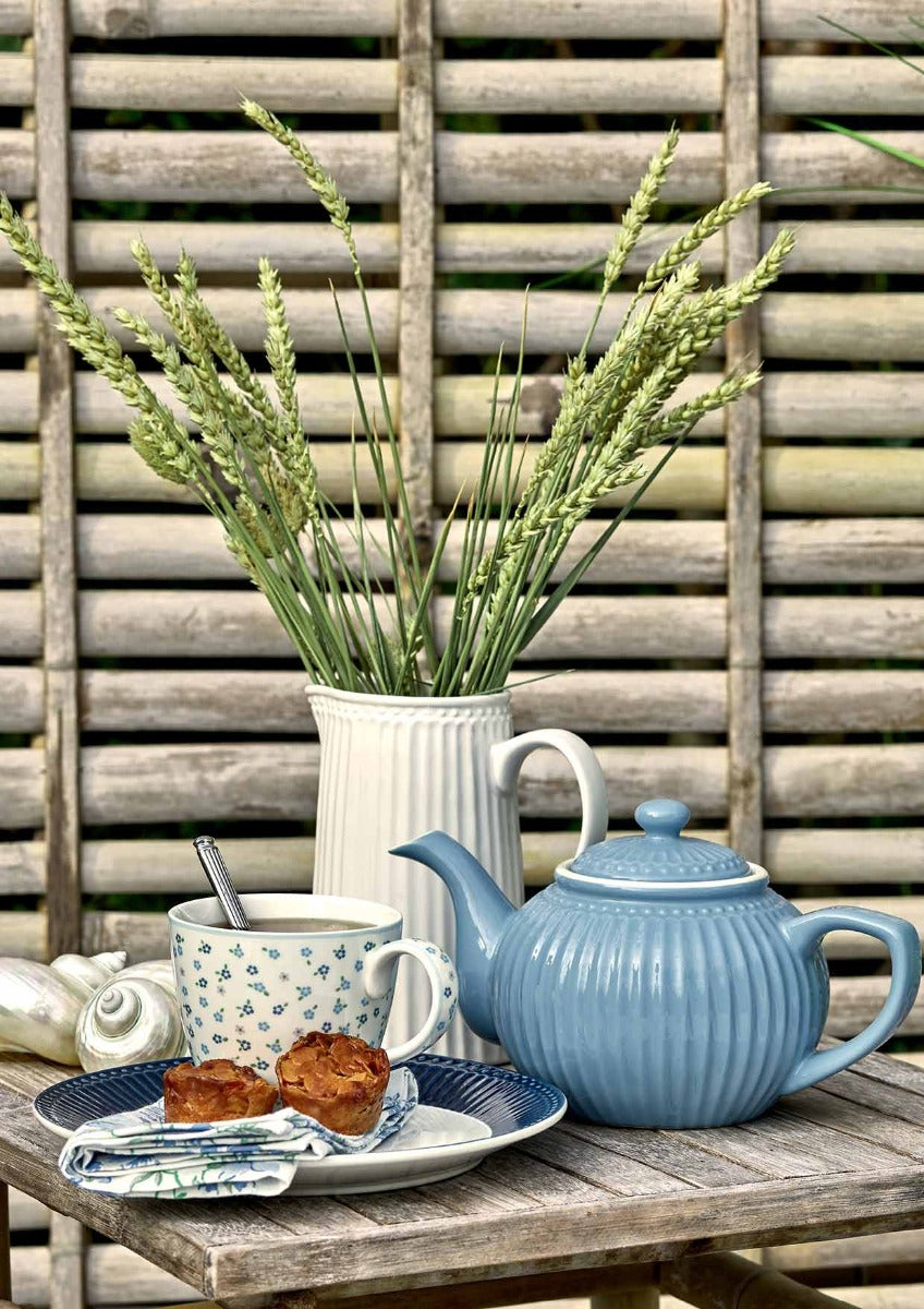 Ein Teeservice mit einer blauen Teekanne, einer weißen GreenGate - Ellise Tasse und Gebäck auf einem kleinen Tisch, neben einer weißen Vase mit grünen Pflanzen, vor einem Holzgitterhintergrund.