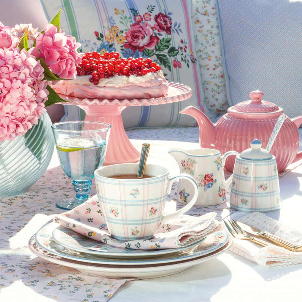 Teegedeck für draußen mit weißen Teetassen, einer Teekanne, einer Tasse Tee und einem mit Beeren garnierten Kuchen aus dem GreenGate - Gaby Besteckkasten auf einem mit Hortensien und einem Blumenkissen geschmückten Tisch.