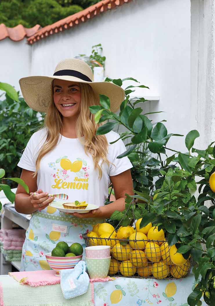 Eine Frau mit einem breitkrempigen Hut und einem Hemd mit Zitronen-Aufdruck lächelt, während sie an einem Gartenstand voller frischer Zitronen eine GreenGate - Pipa Suppenschüssel mehrfarbig hält.