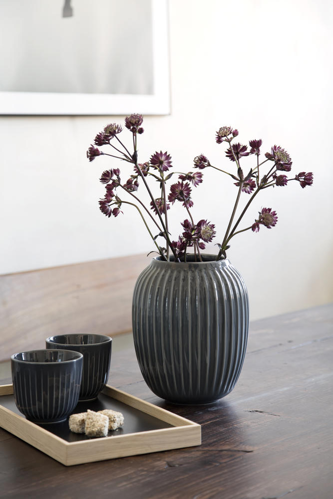 Anthrazitfarbene Vase mit Blüten auf einem Holztisch; daneben zwei dunkle Becher auf einem Holztablett vor heller Wand