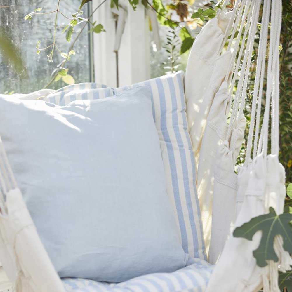 Eine Ib Laursen - Auflage für Stuhl Frederik mit staubig blauen und weissen schmalen Streifen kurz ruht auf einem weißen Hängesessel in einem sonnendurchfluteten Gewächshaus, umgeben von Pflanzen.