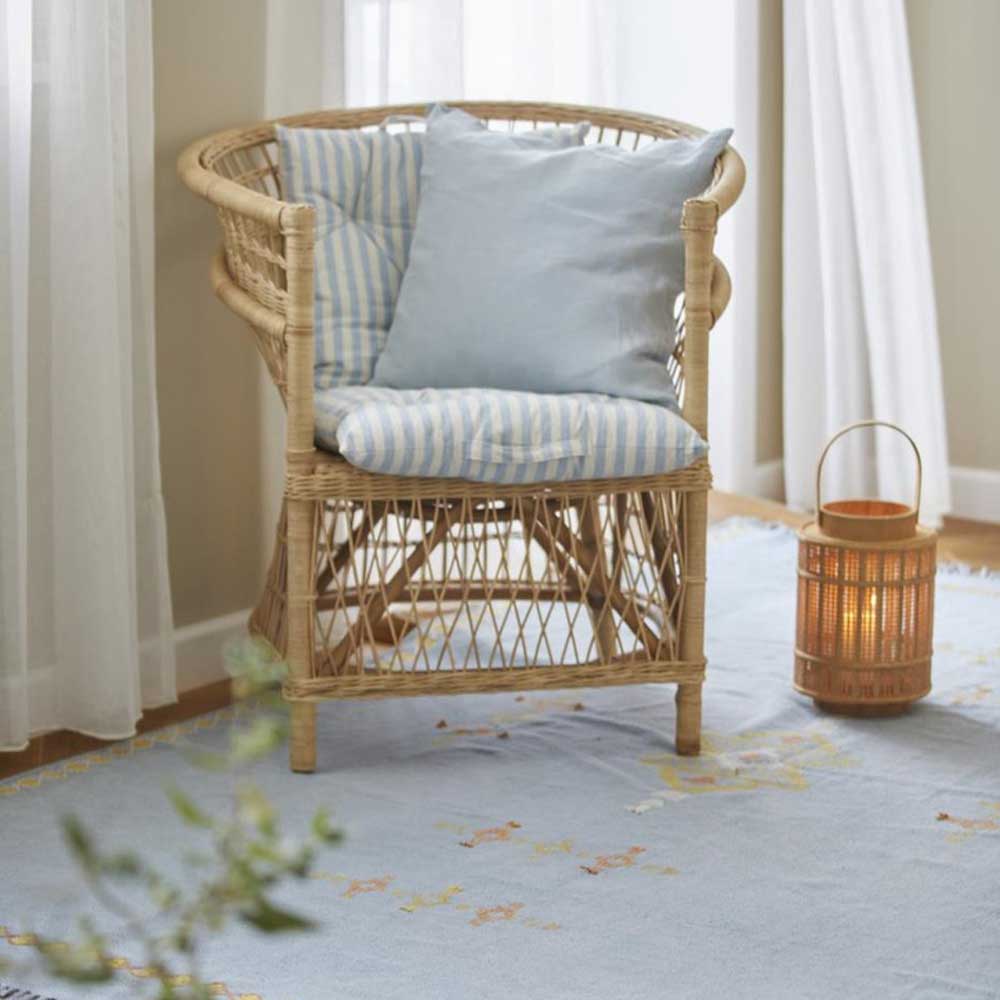 Eine Ib Laursen - Auflage für Stuhl Frederik mit staubig blauen und weissen schmalen Streifen kurz neben einer kleinen Laterne auf einem Teppich mit Blumenmuster in einem sonnendurchfluteten Raum.