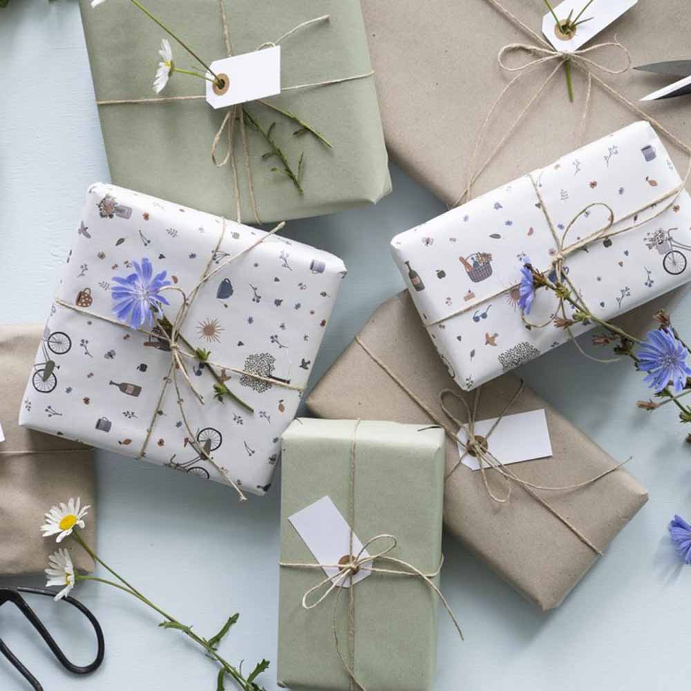Auswahl an wunderschön verpackten Geschenken mit Ib Laursen - Geschenkpapier Sommertage 5 Meter Rolle, Bindfaden und Anhängern, bestreut mit frischen Blumen auf hellblauem Hintergrund.