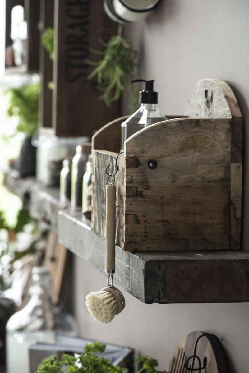 Rustikale Holzregale mit der Ib Laursen - Wandkiste UNIKA bieten Platz für verschiedene Haushaltsgegenstände, darunter eine Reinigungsbürste, Aufbewahrungsbehälter und dekorative Pflanzen in einem neutral getönten Ambiente.