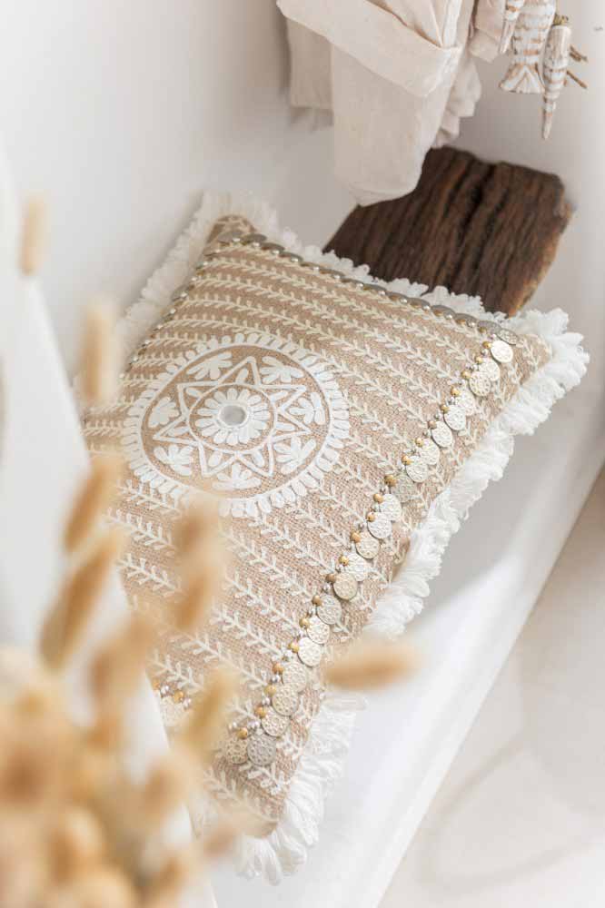 Beiges rechteckiges Kissen mit weißem Kreisornament, Fransen und kleinen goldfarbenen Plättchen auf einer hellen Sitzfläche vor dunklem Holz.