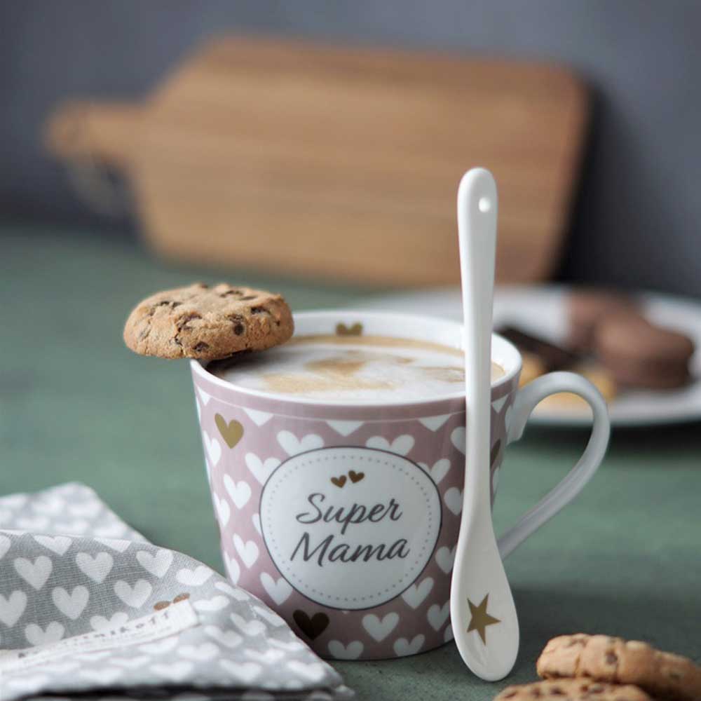 Ein Krasilnikoff - Happy Cup „Super Mama Hearts“ mit einem Löffel und einem Keks darauf, auf einem Tisch mit einem weiteren Teller und Keksen im Hintergrund.