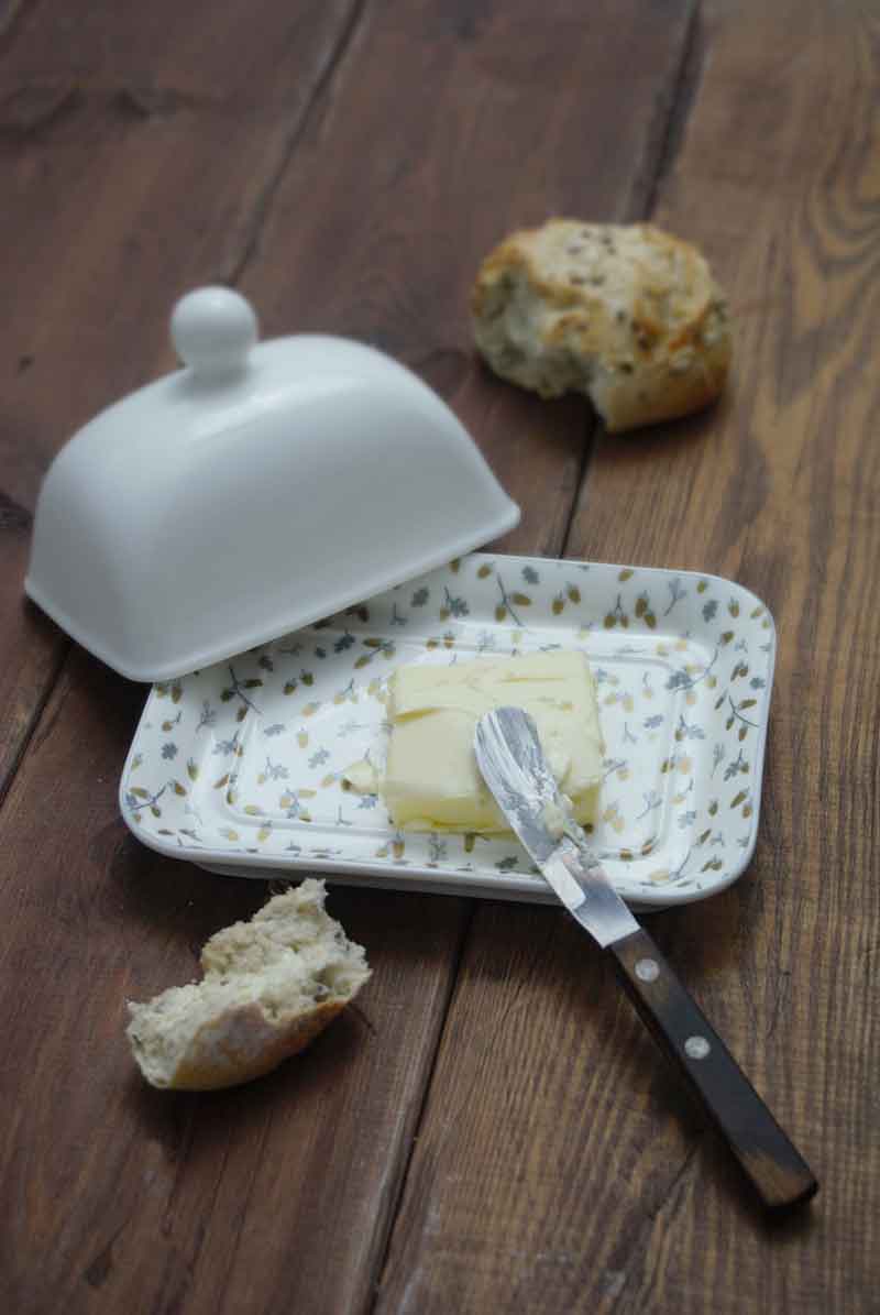 Eine Krasilnikoff-Butterdose mit Deckel in Weiß, darin ein Stück Butter und ein Buttermesser, neben einem halb aufgegessenen Brötchen auf einem Holztisch.