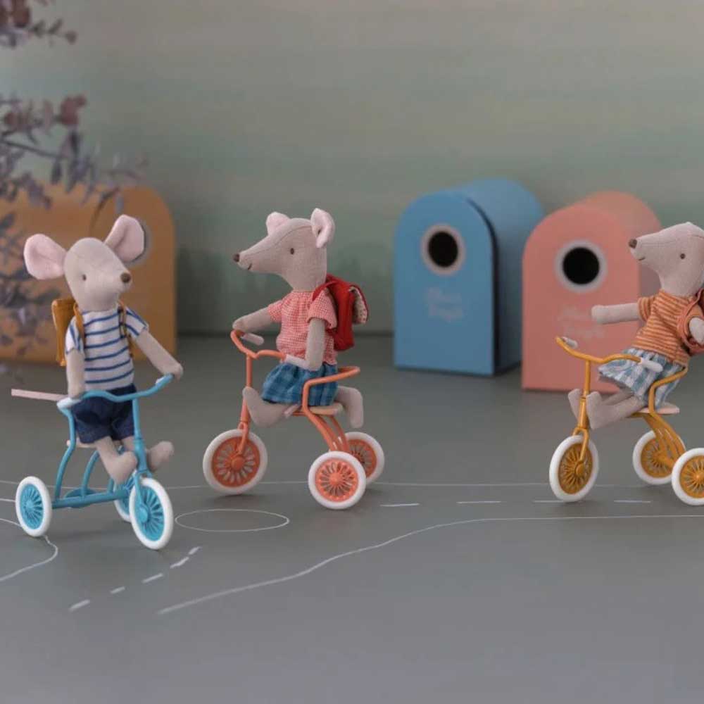 Drei Maileg-Maus „Big Sister“ – Dreiradmaus mit Tasche fahren auf Dreirädern auf einem gezeichneten Weg, mit farbenfrohen Blockhintergründen in sanft gefärbter Umgebung.