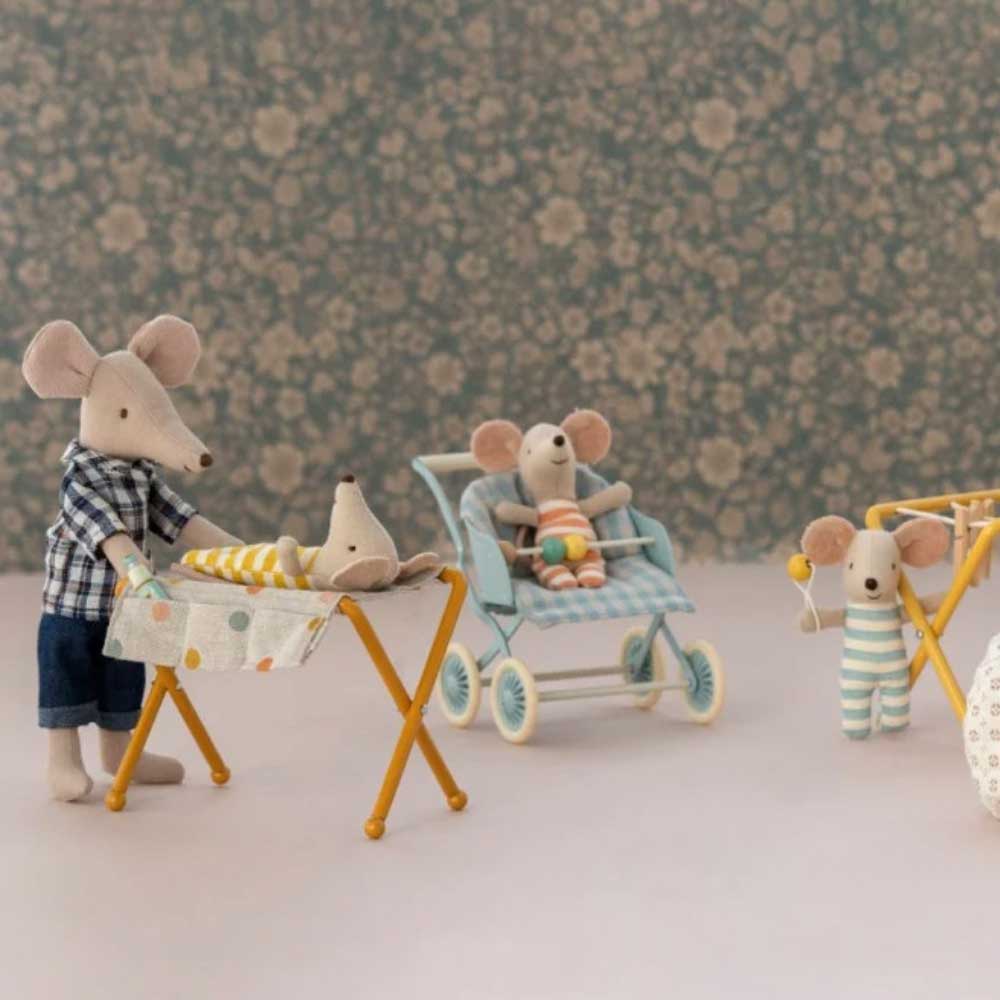 Angezogene Maileg-Spielzeugmäuse interagieren mit Miniatur-Requisiten wie einem Kinderwagen und einem Maileg - Puppenhaus Wickeltisch für Maus vor einem floralen Hintergrund.