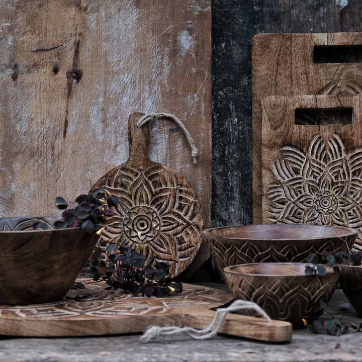 Rustikales Küchengeschirr aus Holz von Majas Cottage mit aufwendigen Schnitzereien auf einem verwitterten Tisch, darunter Schüsseln, Schneidebretter und ein mit einem Blumenmuster verzierter Dekorationsgegenstand.